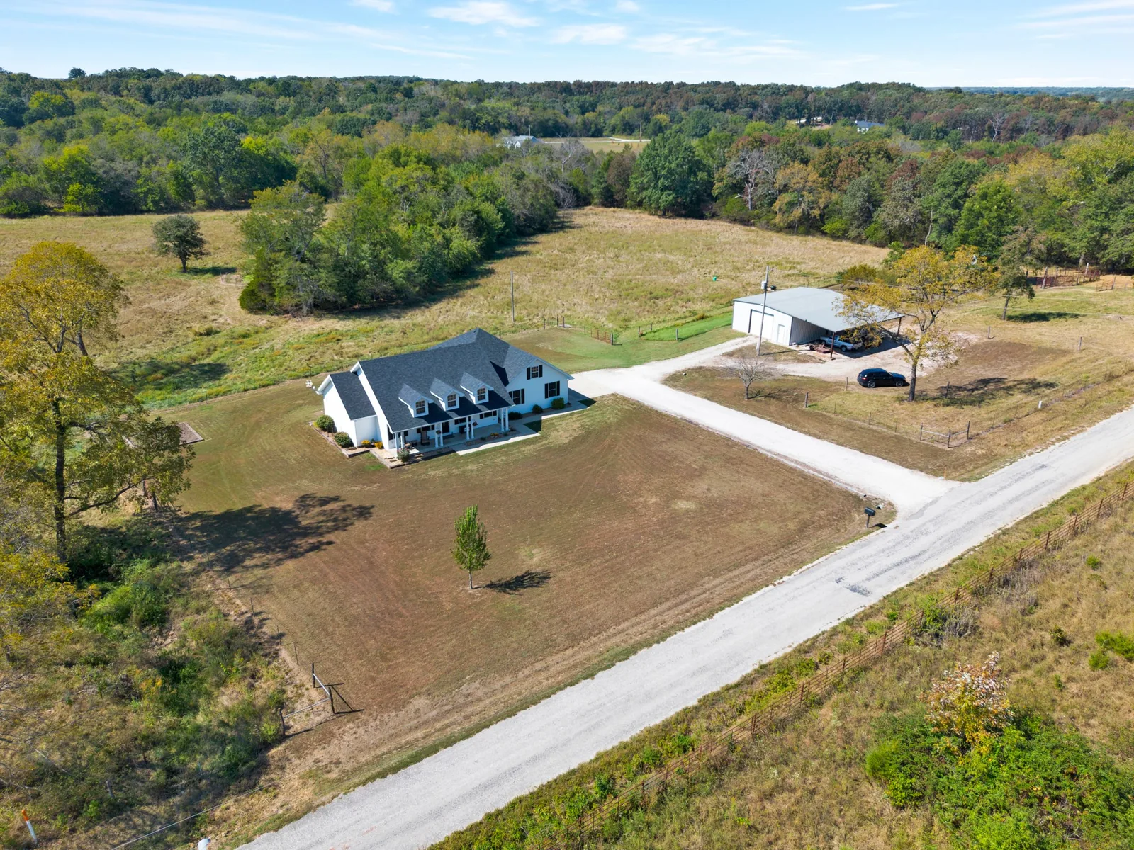 Aerial view of the white country home, a gravel drive and a metal shop building with pasture behind