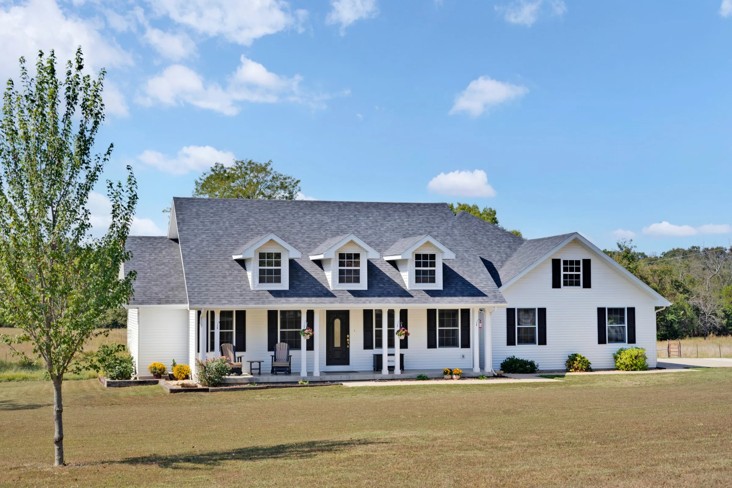 White Cape Cod style home with three dormers, black shutters and a covered front porch on a wide lawn