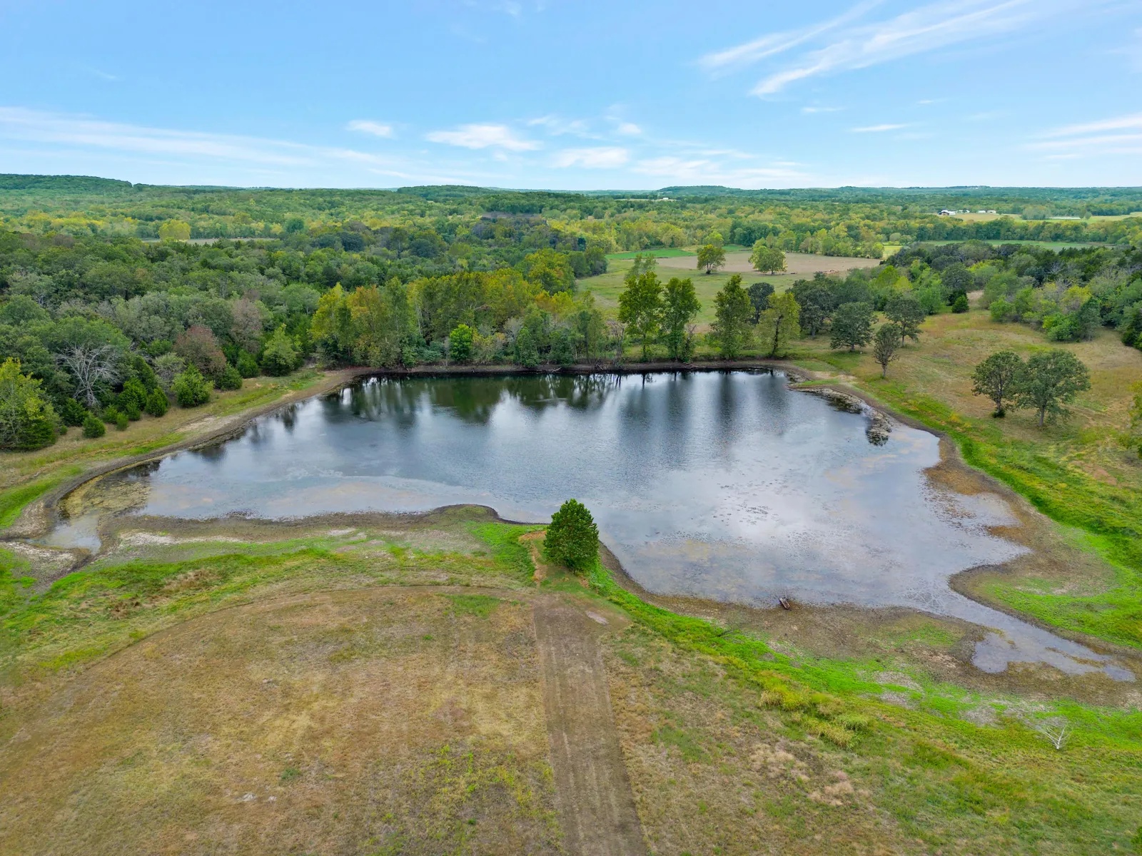 Aerial view of a pond on open acreage with wooded hills beyond